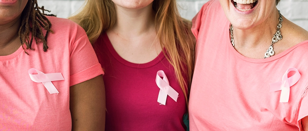 Three women sit and smile while wearing pink shirts and breast cancer ribbons.